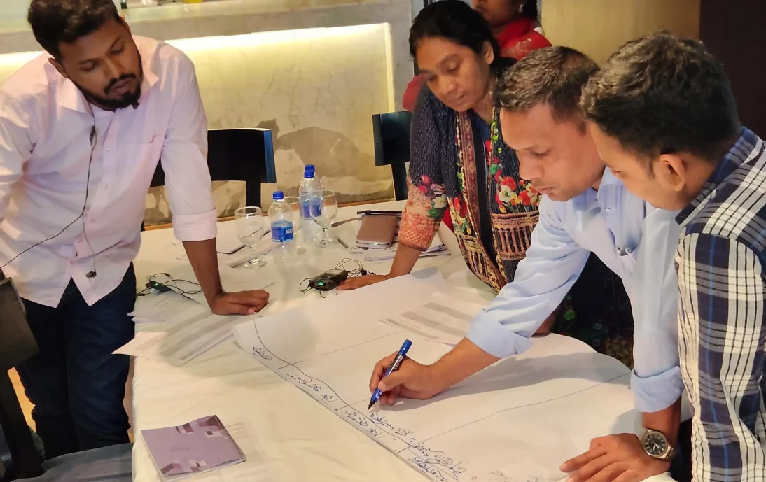 Data analysis methods training in Bangladesh: A group of people stood around a table, writing on a large sheet of paper.