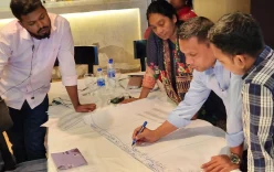 Data analysis methods training in Bangladesh: A group of people stood around a table, writing on a large sheet of paper.